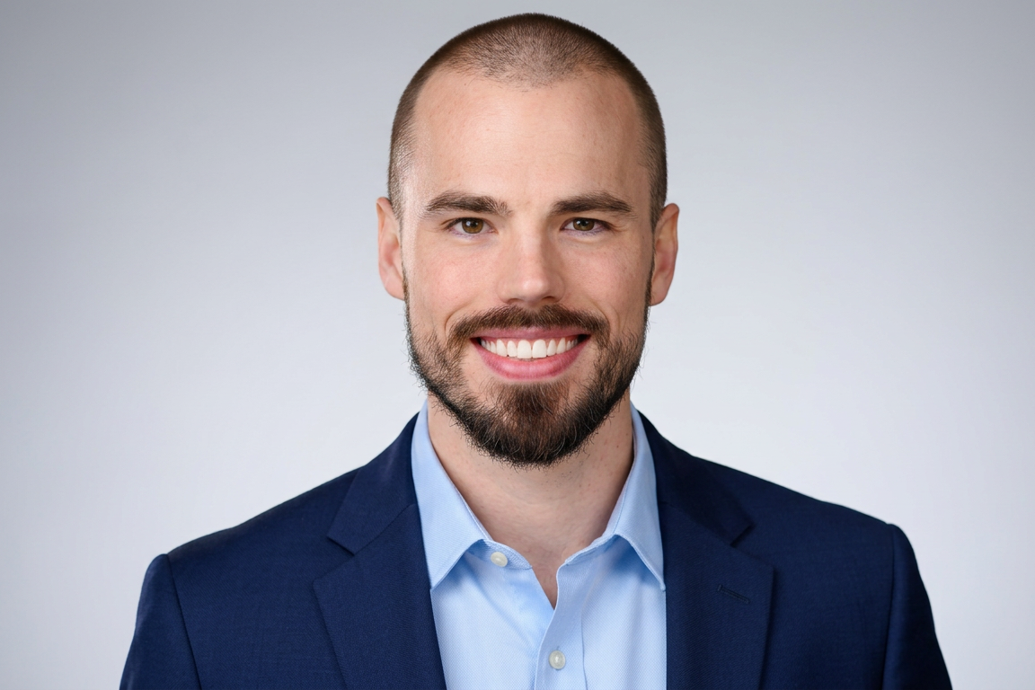 A smiling man with a short beard and a shaved head wearing a blue suit and light blue shirt against a neutral background.